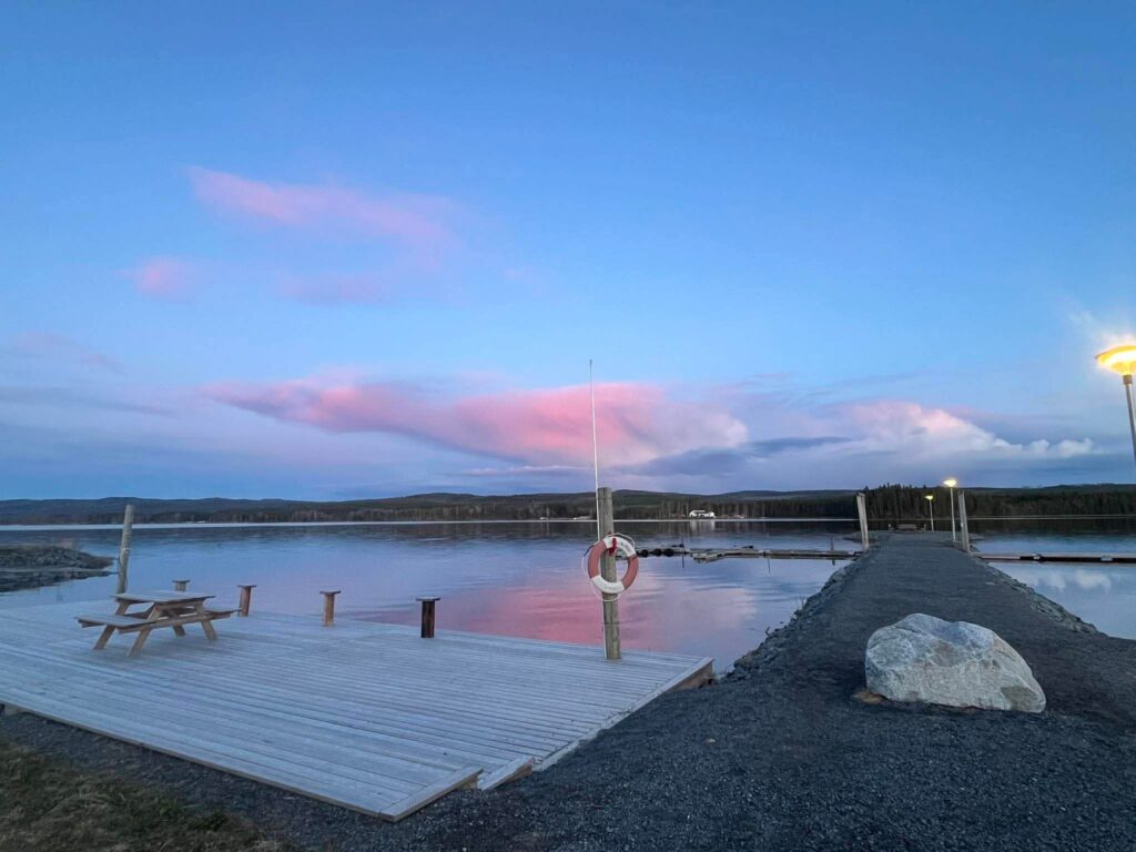 Picknickbord på trädäck vid sjö, lyktor och livboj, rosa skymningshimmel speglas i vattnet