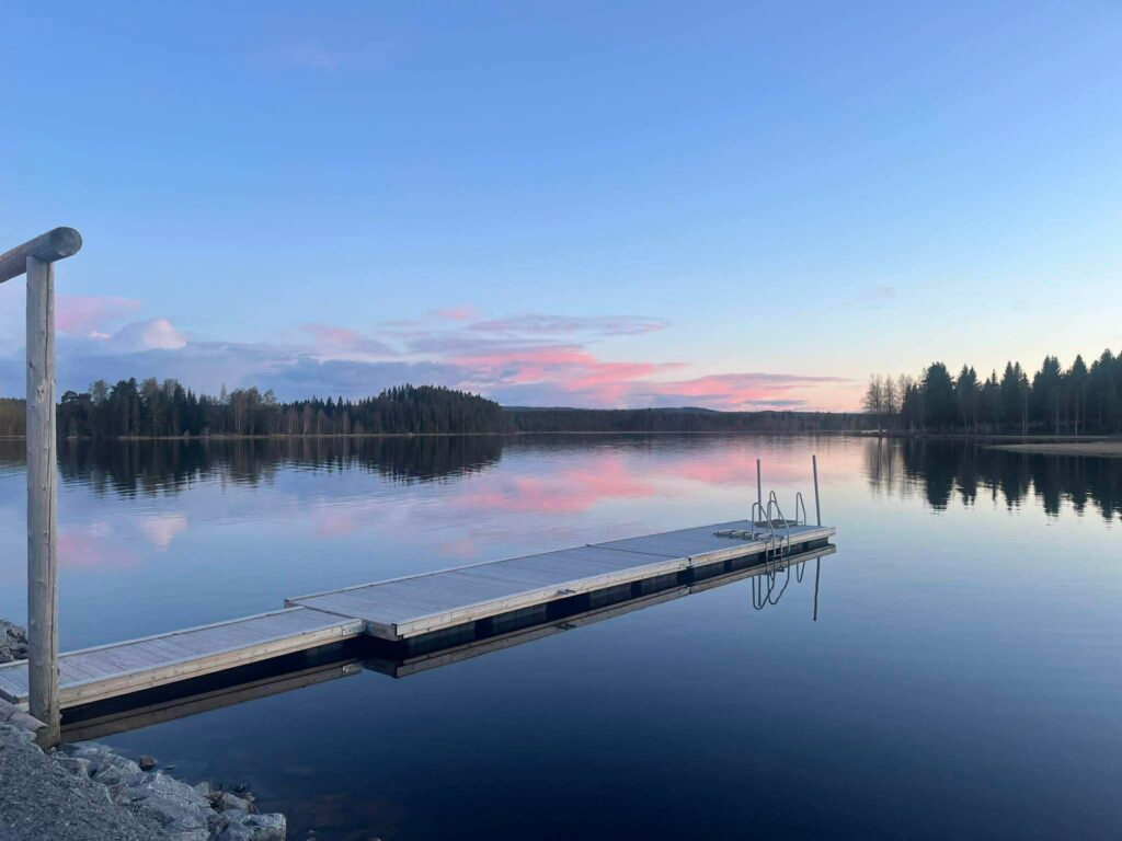 Träbrygga sträcker sig ut i blank sjö, rosa moln speglar sig i vattnet vid solnedgång