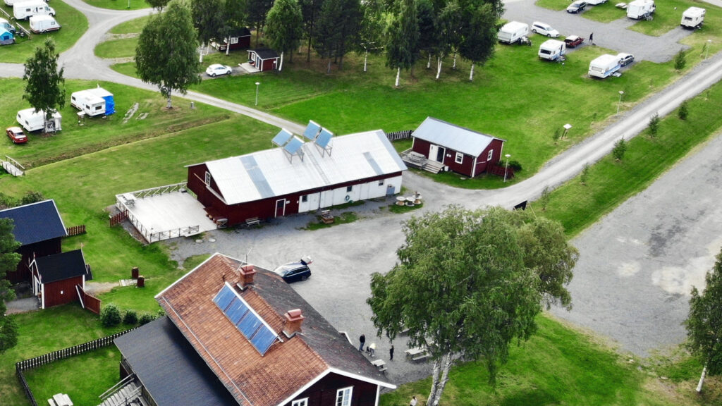 Flygbild över Ånge Camping. Det långa röda huset med vit gavel ligger mitt i bild, det lilla röda huset strax till höger om det, och husvagnarna står på gräsytorna runt omkring.